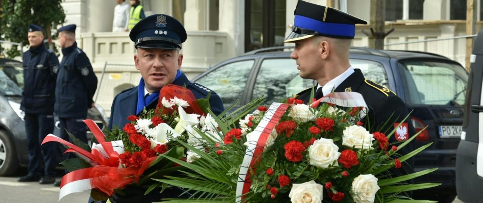 policjant i strażak trzymają wieńce biało czerwone, w tle budynek i pojazdy 