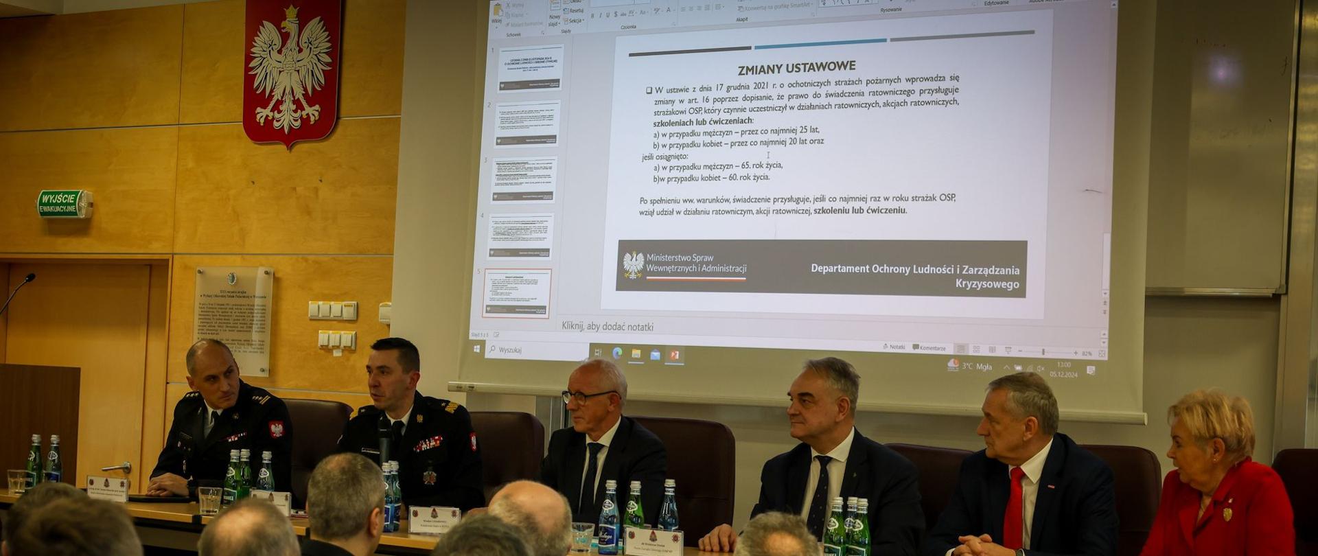 Sześć osób siedzi za stołem prezydialnym, za nimi na ekranie widoczny napis "zmiany ustawowe" dotyczący ustawy o ochotniczych strażach pożarnych. Na pierwszym planie widać osoby w mundurach OSP i PSP słuchające wypowiedzi osób za stołem. Za stołem siedzą w czarnych mundurach wyjściowych komendant główny PSP, rektor-komendant akademii Pożarniczej, sekretarz stanu i podsekretarz stany w MSWiA, prezes Zarządu Głównego Związku OSP RP oraz przewodnicząca Sejmowego Zespołu Strażaków.