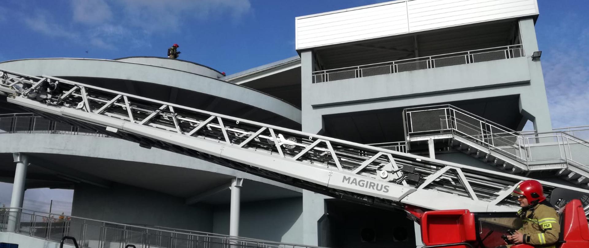 boczne ujęcie drabiny pożarniczej rozkładającej się do dachu budynku, operator siedzący w miejscu operatora sprzętu. W tle budynku wielopoziomowy garaż ze spiralą do zjazdu oraz na dachu obiektu znajdujący się strażak
