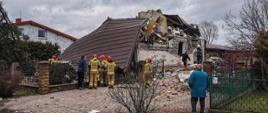 Na zdjęciu strażacy w akcji w ubraniach koloru musztardowego na tle budynku zawalonego w wyniku wybuchu gazu, wokół porozrzucane elementy budynku, dach wyrzucony poza budynek.