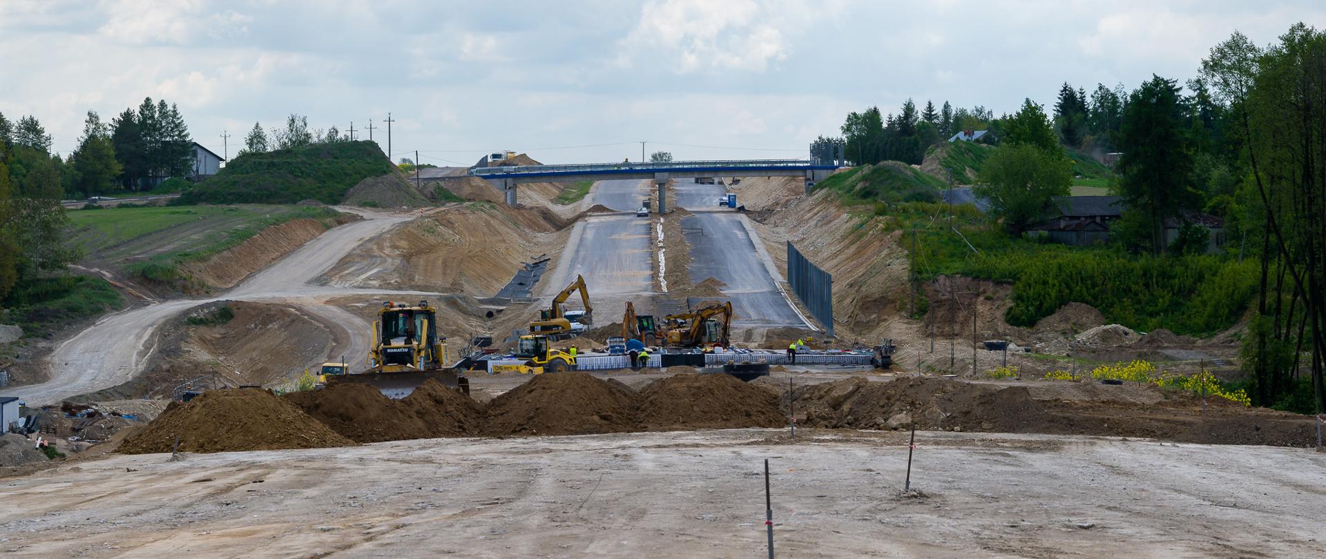 Budowa drogi ekspresowej w zaawansowanym etapie – widok na szeroki pas ziemi przygotowany pod nawierzchnię, z maszynami budowlanymi (koparki, spycharki) pracującymi w tle. Po obu stronach widoczne skarpy i ekrany akustyczne, a w oddali wiadukt nad przyszłą trasą. Otoczenie to tereny zielone z drzewami i zabudową w tle.