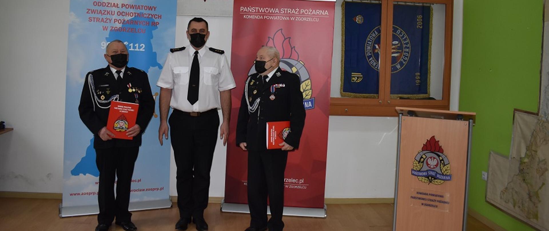 Widok na część pomieszczenia, podłoga wyłożona panelami w jasnym kolorze. Na wprost biała ściana, na której wisi godło państwowe. Przy ścianie stoją banery. Od lewej w kolorze błękitnym i logiem ZOSP RP, Z prawej w kolorze bordowym i logiem PSP. Za banerem PSP na ścianie wisi gablota, w której umieszczony jest sztandar. KP PSP w Zgorzelcu. Ściana z prawej strony jest w kolorze zielonym. Wiszą na niej dwie mapy obrazujące powiat zgorzelecki. Na tle banerów stoi funkcjonariusz PSP ubrany w białą koszulę, krawat i czarne spodnie. Na twarzy ma czarną maseczkę ochronną. Po jego lewej i prawej stronie stoją druhowie OSP ubranych w mundury galowe ze sznurem galowym w kolorze srebrnym oraz czarne maseczki ochronne. Na mundurach druhów wdać dużo medali. Obaj druhowie trzymają w rękach teczki papierowe w kolorze czerwony, na których widać logo PSP.