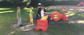 Na zdjęciu widzimy strażaka oraz 3 dzieci na placu zabaw. Jeden z chłopków leje wodę z węża strażackiego