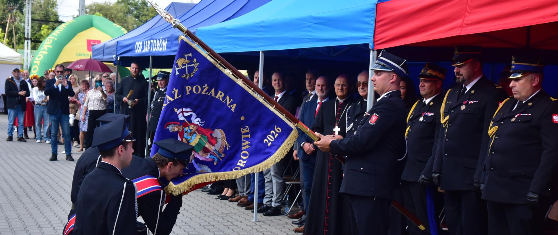 Podczas obchodów 100-lecia OSP Jaktorów strażacy klęczą przed nowym sztandarem jednostki. W uroczystości uczestniczy Mazowiecki Komendant Wojewódzki PSP nadbryg. Artur Gonera, stojący wśród zaproszonych gości i przedstawicieli straży pożarnej podczas ceremonii związanej z przekazaniem sztandaru.