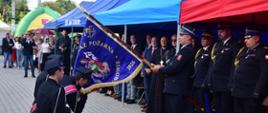 Podczas obchodów 100-lecia OSP Jaktorów strażacy klęczą przed nowym sztandarem jednostki. W uroczystości uczestniczy Mazowiecki Komendant Wojewódzki PSP nadbryg. Artur Gonera, stojący wśród zaproszonych gości i przedstawicieli straży pożarnej podczas ceremonii związanej z przekazaniem sztandaru.