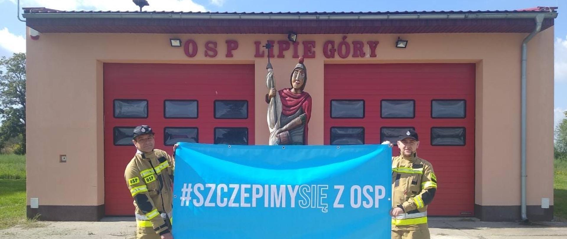 Fotografia przedstawia dwóch strażaków - ochotników w ubraniach specjalnych trzymających rozciągnięty baner akcji #Szczepimy się z OSP. Strażacy stoją przed jednostką OSP Lipie Góry. W tle budynek i bramy garażowe.
