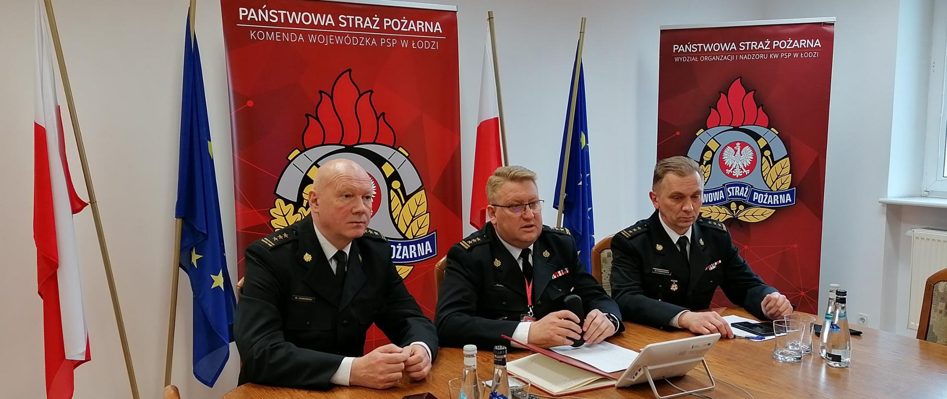 Na zdjęciu widać trzech komendantów wojewódzkich PSP w Łodzi w czasie wideokonferencji, obrani są w mundury wyjściowe w tle dwa roll-upy komendy wojewódzkiej PSP w Łodzi, 2 flagi polski i 2 unii europejskiej