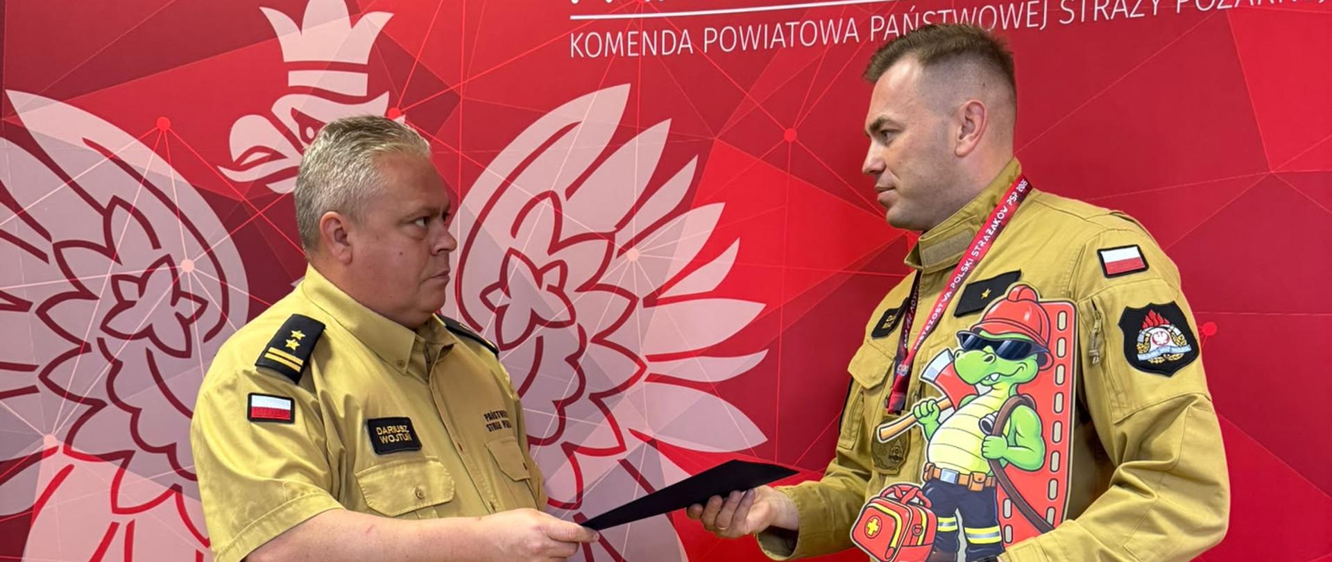 Komendant Powiatowy PSP wręcza wyróżnienie funkcjonariuszowi. Strażacy w piaskowych mundurach podają sobie dłonie na tle ścianki Państwowej Straży Pożarnej.