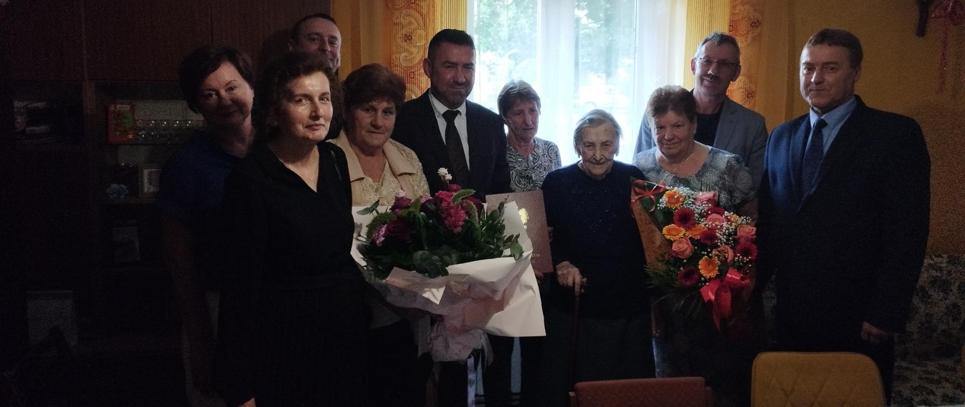 Grupa osób pozuje do zdjęcia w pokoju. Pośrodku stoi starsza pani z laską. Dwie kobiety stojące po jej obu stronach trzymają dwa duże bukiety kwiatów, a stojący obok mężczyzna trzyma dyplom w ozdobnej teczce.