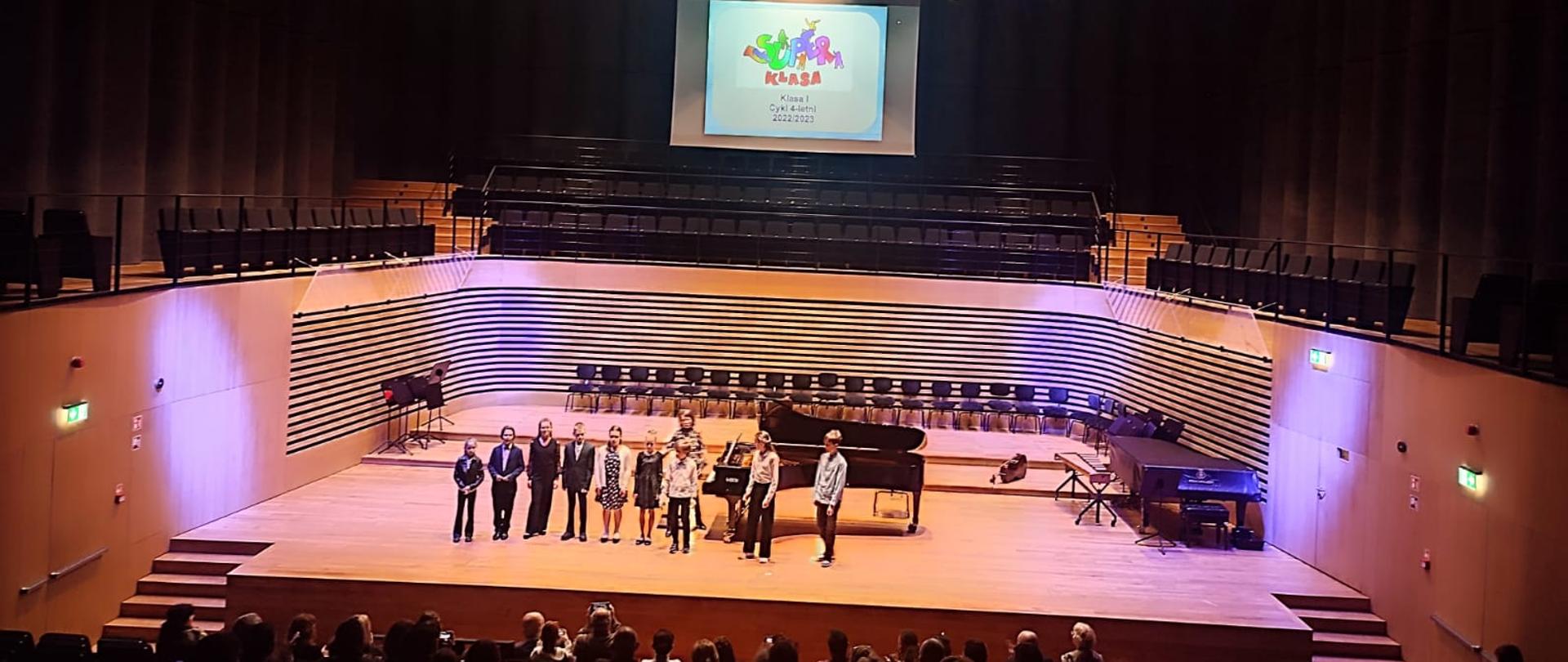 Dziewięcioro dzieci stoi w rzędzie na scenie, za nimi kobieta i fortepian. Nad scena widać rzutnik z kolorowym napisem Super klasa - klasa I cykl 4-letni 2022/2023.