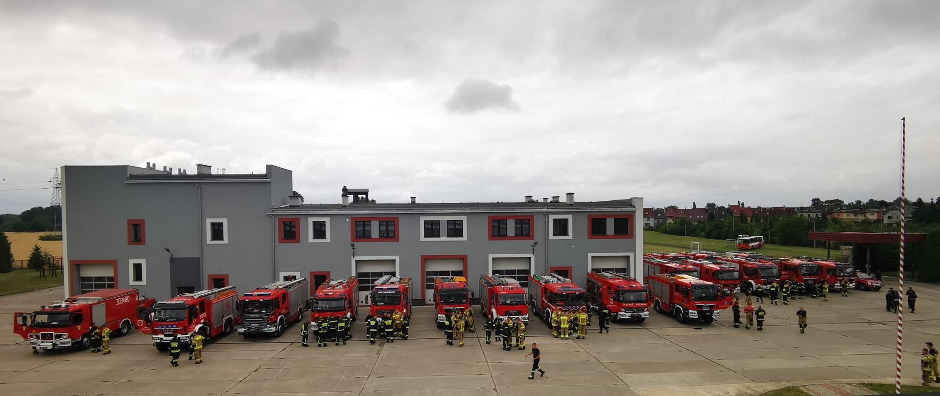 20 wozów strażackich stojących w rzędzie przy budynku straży pożarnej. Obok wozów stoją zastępy strażaków.