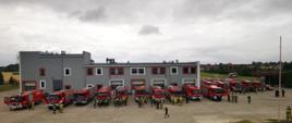 20 wozów strażackich stojących w rzędzie przy budynku straży pożarnej. Obok wozów stoją zastępy strażaków.