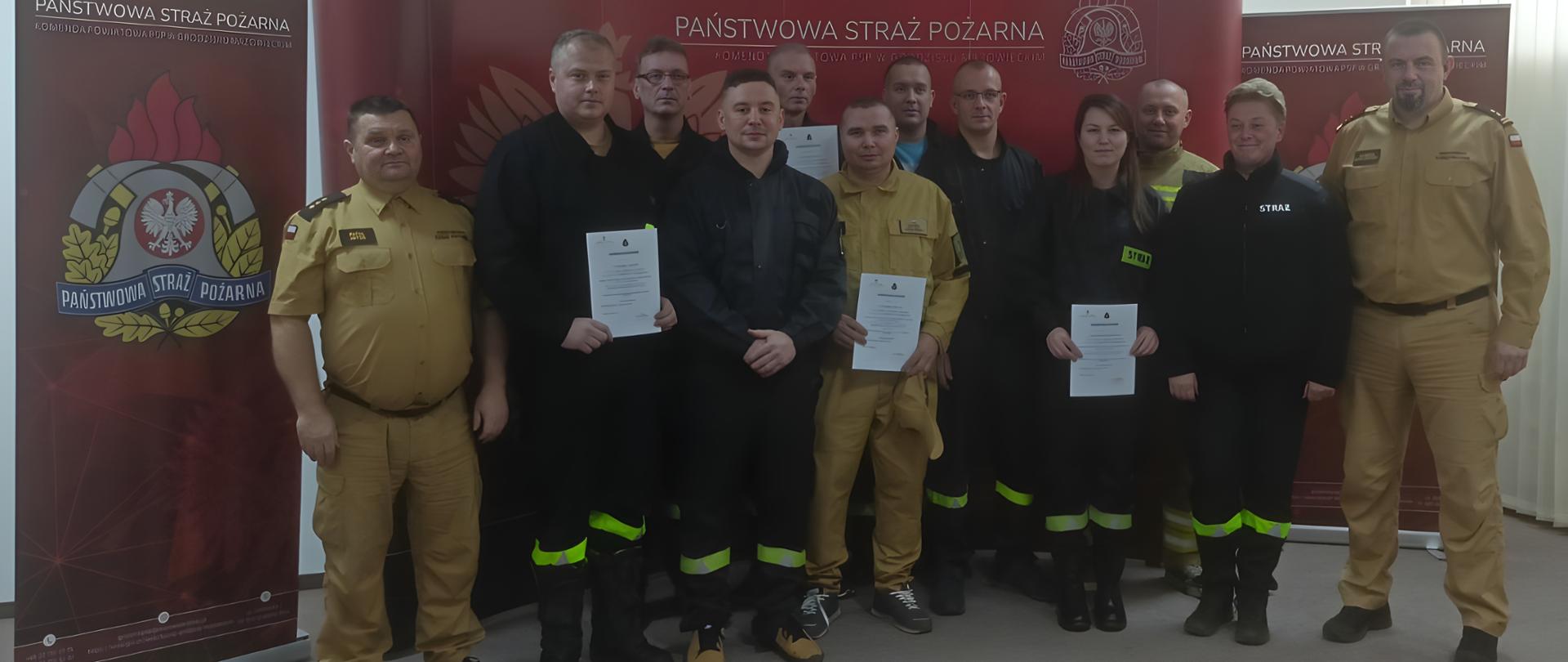 Zdjęcie grupowe przedstawiające dwunastu strażaków (dziesięciu mężczyzn i dwie kobiety) w mundurach służbowych, stojących przed czerwonym tłem z logiem i napisem "PAŃSTWOWA STRAŻ POŻARNA". Kilka osób trzyma w rękach certyfikaty lub dyplomy.