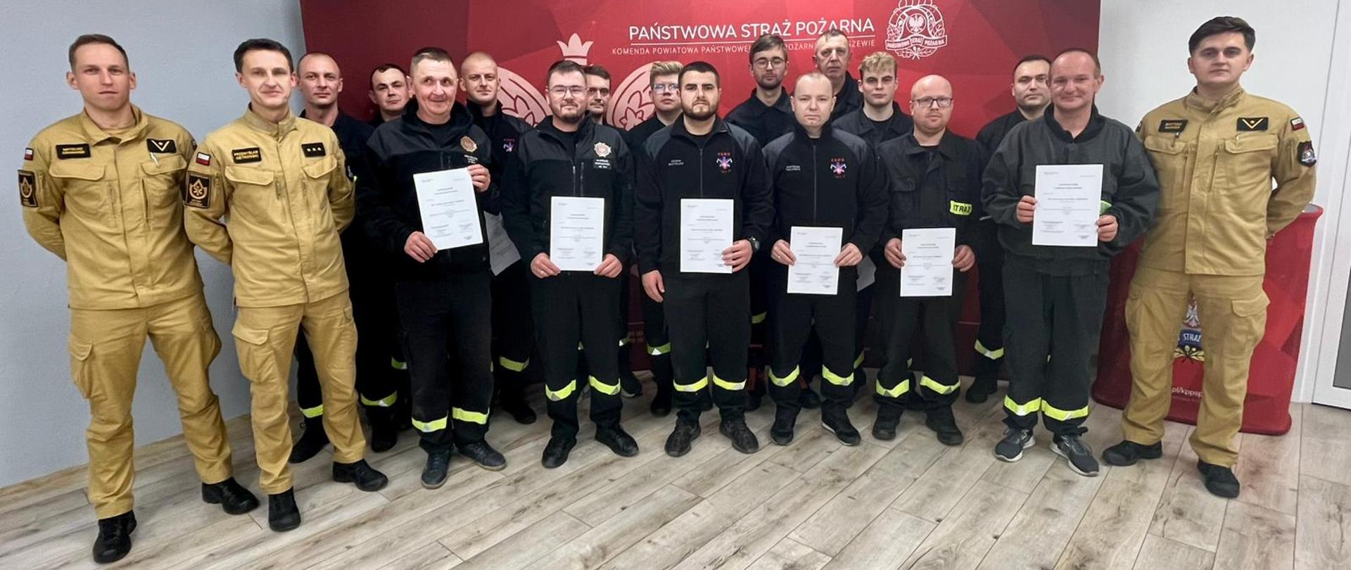 grupa osób ubrana w mundury strażackie na tle czerwonej ścianki z zaświadczeniami w rękach