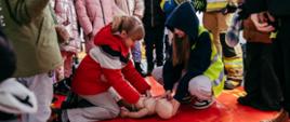 Dzieci wykonują resuscytację krążeniowo-oddechową na fantomie szkoleniowym pod okiem strażaków podczas pokazów ratownictwa lodowego, w tle dzieci oraz strażacy