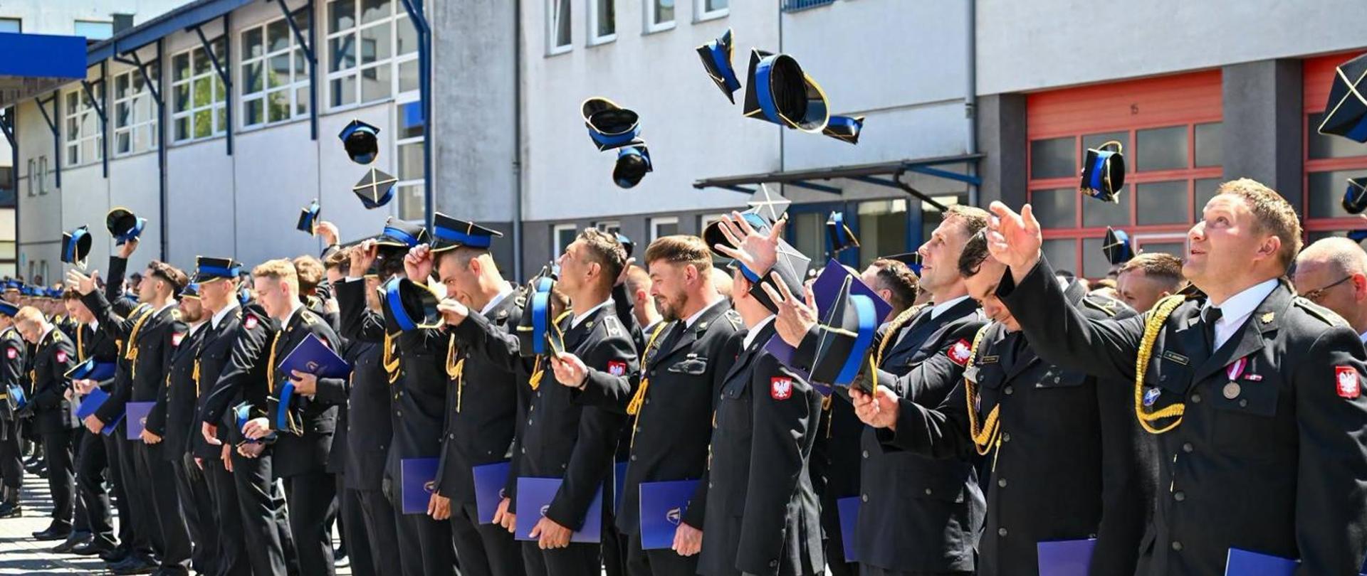 Uroczystą promocja absolwentów Szkoły Aspirantów Państwowej Straży Pożarnej w Poznaniu. Grupa kilkudziesięciu nowo promowanych strażaków Państwowej Straży Pożarnej stoi podczas uroczystego apelu na placu przed budynkiem szkoły. Z radością podrzucają w górę czapki rogatywki podczas uroczystej promocji, w rękach trzymają dyplomy. W tle widać budynek jednostki.
