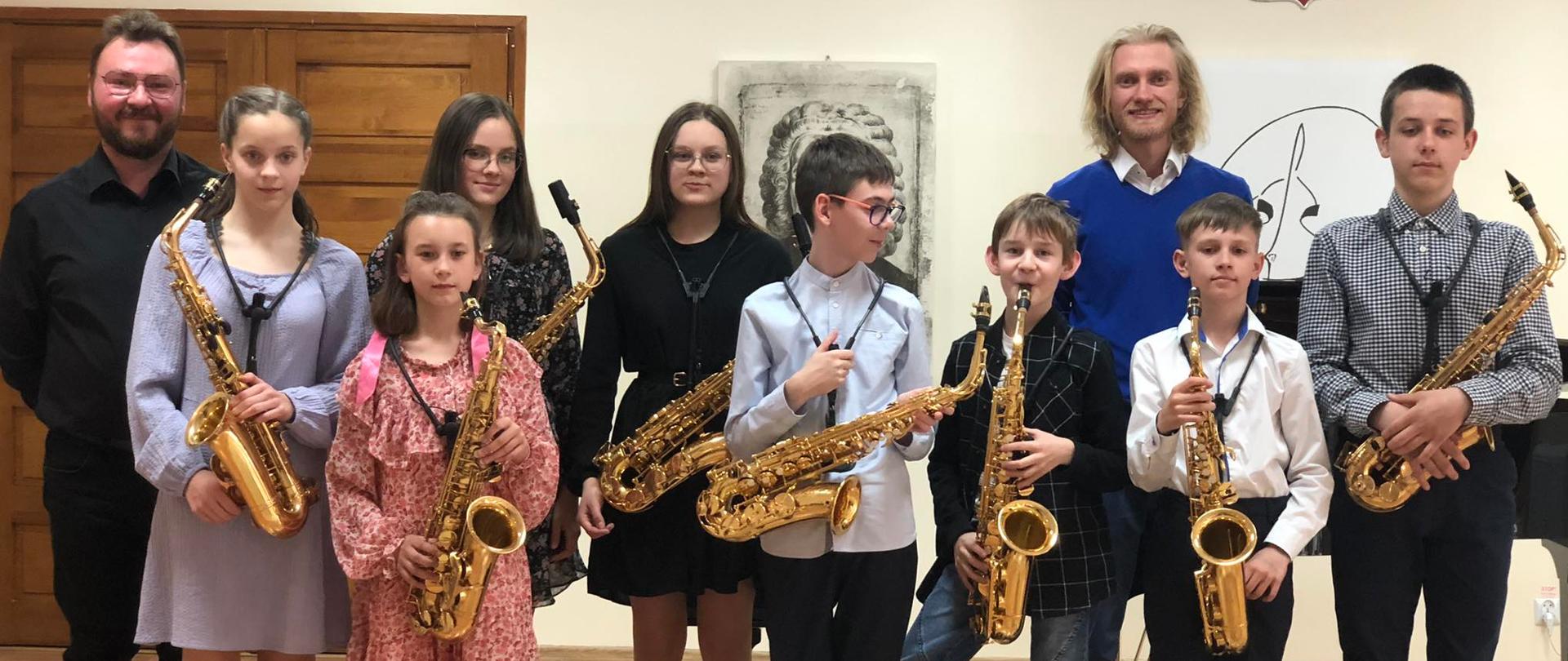 Ośmioro odświętnie ubranych dzieci, cztery dziewczynki i czterech chłopców stoi na scenie i trzyma złote saksofony. Obok nich stoi ich nauczyciel ubrany na niebiesko oraz akompaniator ubrany na czarno 