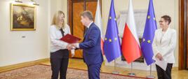 Minister Jan Grabiec wręcza akt powołania do Rady KSAP dr Karolinie Kulickiej, obok stoi szefowa służby cywilnej Anita Noskowska-Piątkowska; w tle flagi Polski i Unii Europejskiej
