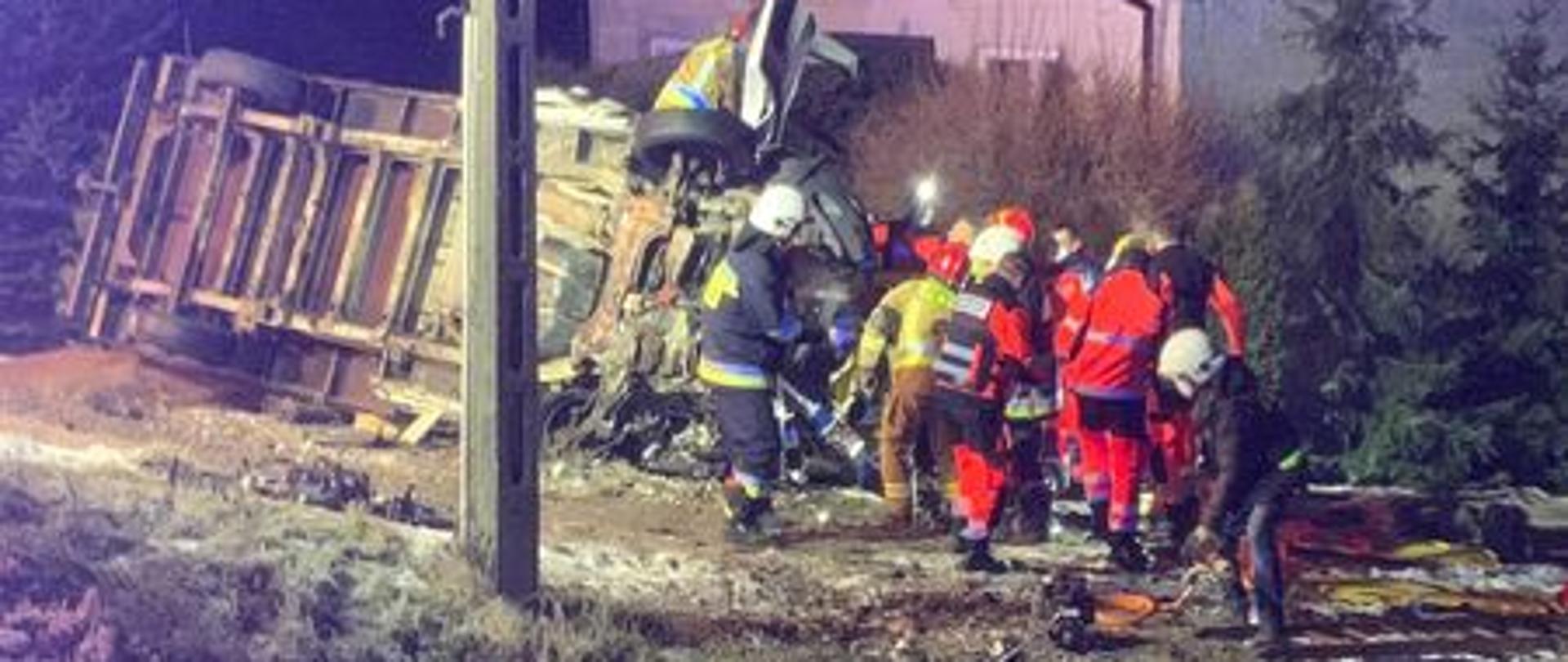 Samochód dostawczy prze2wrocony na boku ,leży poza jezdnią. Strażacy Jednostek PSP I OSP oraz ratownicy medyczni wykonują dostęp osób znajdujących się w pojeździe.