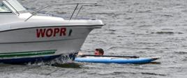 Mężczyzna znajdujący się w wodzie. Obok mężczyzny znajduje się deska SUP oraz wiosło. Do mężczyzny podpływa łódź z czerwonym napisem WOPR na boku łodzi.