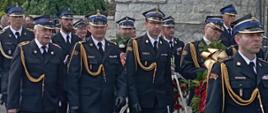 Uczestnicy pogrzebu w przeważającej większości w mundurach strażackich idą w kondukcie pogrzebowym. W tle zabudowania miejskie.