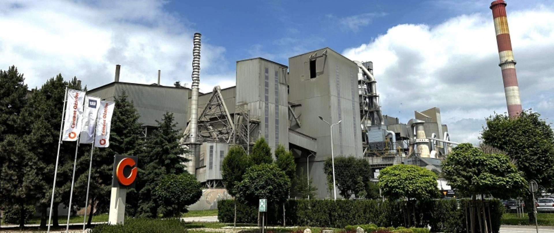 Zakład przemysłowy z flagami, kominami i budynkami, otoczony drzewami i krzewami, pod częściowo zachmurzonym niebem.