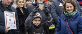 W środkowej części zdjęcia odznaczony chłopiec podnoszący nad głowę trzymaną w dłoniach odznakę. Z lewej tata Marcina, z prawej mama, za chłopcem dyrektor jego szkoły, z prawej za mamą burmistrz Koronowa.