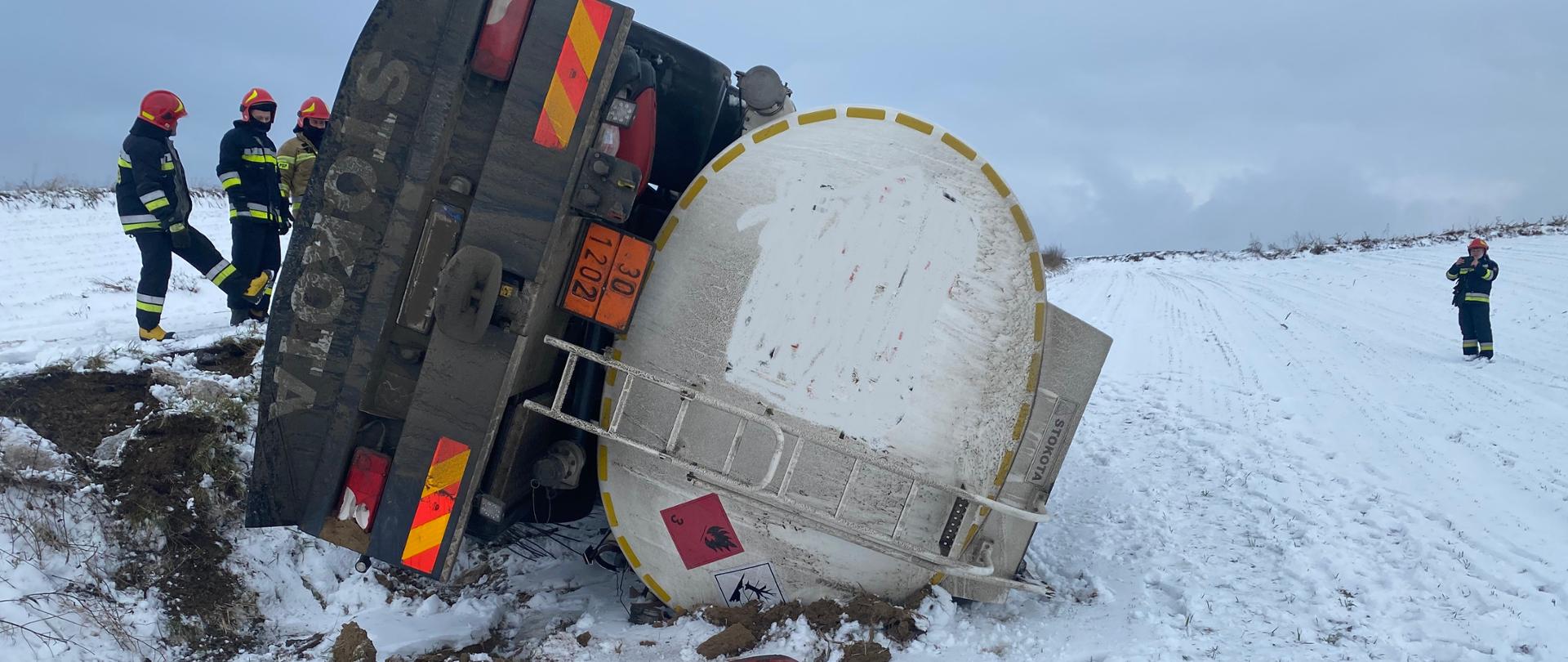 Przewrócona na bok cysterna paliwowa, obok znajdują się strażacy, którzy zabezpieczają teren działań.
