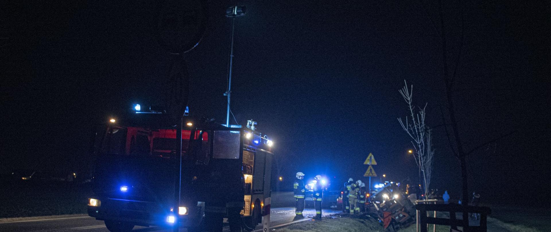 Zdjęcie przedstawia samochód strażacki, który oświetla teren prowadzonych działań. W dalszym planie ciągnik rolniczy leżący na boku w rowie, strażacy wykonujący czynności służbowe. 