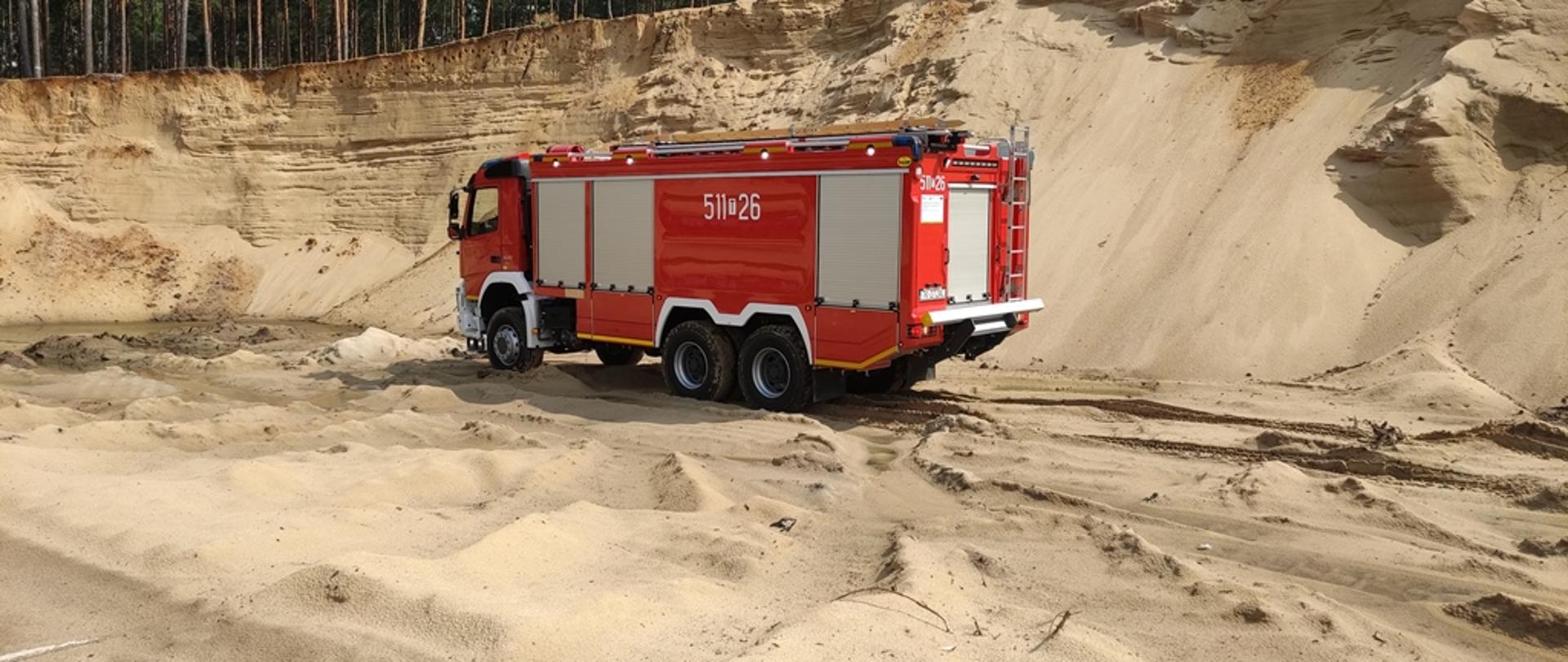 Teren piaskowni, na pierwszym planie pojazd pożarniczy poruszający się po wyrobisku po wydobyciu piasku, na drugim planie skarpy piasku