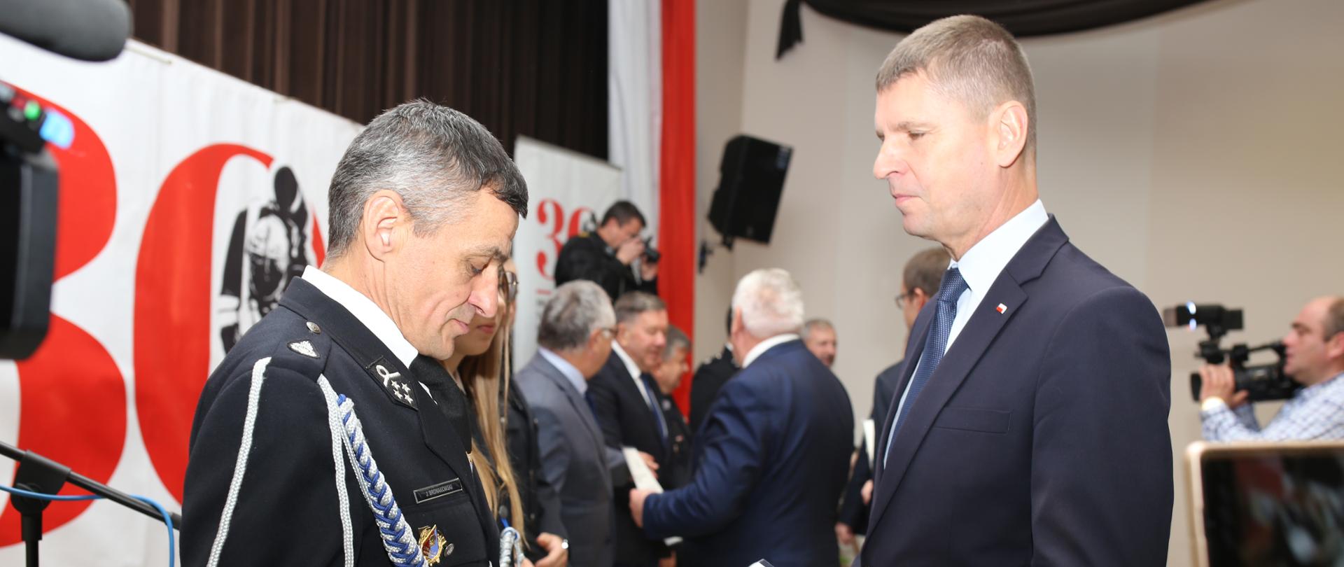 Dariusz Piontkowski - Sekretarz Stanu Ministerstwa Edukacji i Nauki przekazuje promesy druhom OSP. w tle banery związane ze strażą pożarną.