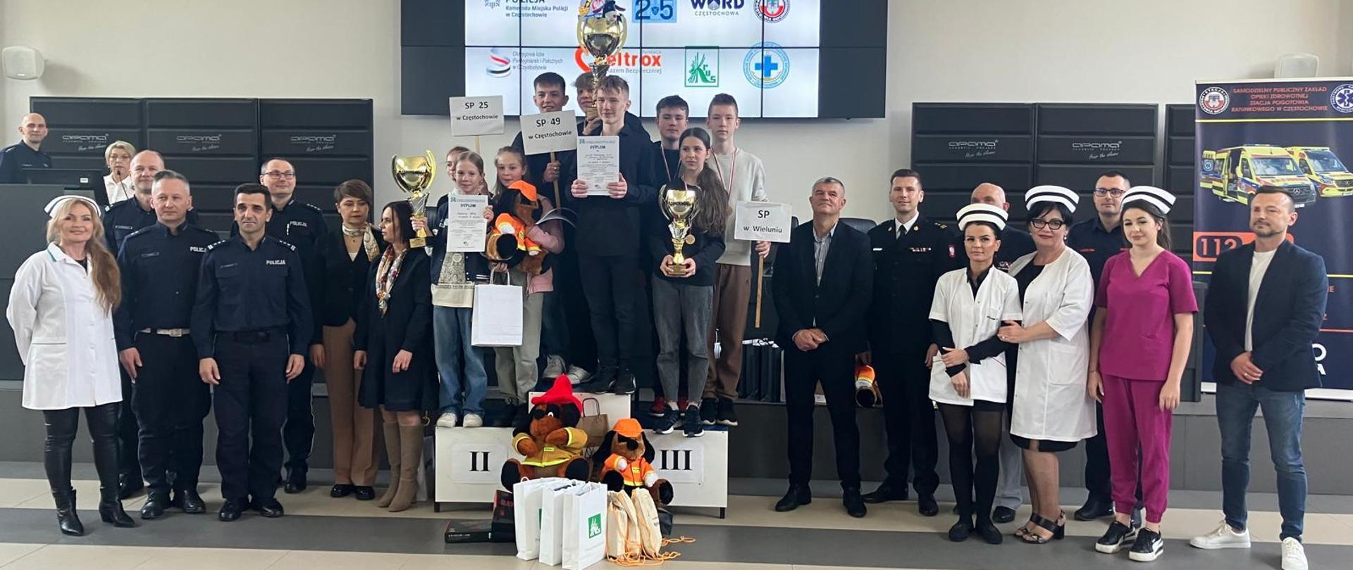 Zdjęcie grupowe laureatów konkursu, którzy stoją na podium trzymając w rękach zdobyte puchary. Wokół nich jurorzy oraz przedstawiciele Policji, Państwowej Straży Pożarnej, Miasta Częstochowy oraz Służby Medycznej. W tle widać telebim, na którym wyświetlana jest nazwa konkursu.
