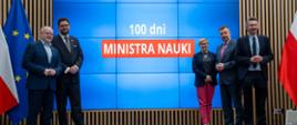 Minister Wieczorek i wiceministrowie stoją na sali, pod wielkim wbudowanym w ścianę ekranem z napisem 100 dni ministra nauki.