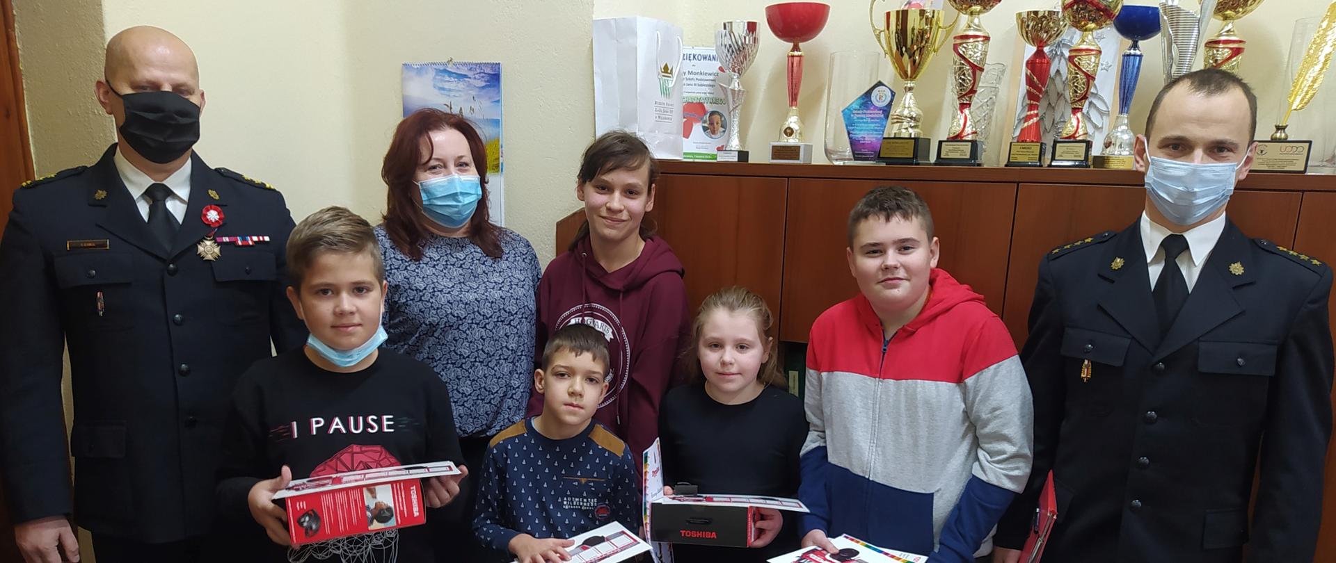 Pięcioro dzieci stoi w pomieszczeniu. Za nimi stoi pani dyrektor szkoły. Z lewej strony stoi komendant powiatowy PSP w Żyrardowie, a z prawej strony rzecznik prasowy komendanta.