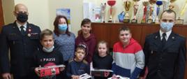 Pięcioro dzieci stoi w pomieszczeniu. Za nimi stoi pani dyrektor szkoły. Z lewej strony stoi komendant powiatowy PSP w Żyrardowie, a z prawej strony rzecznik prasowy komendanta.