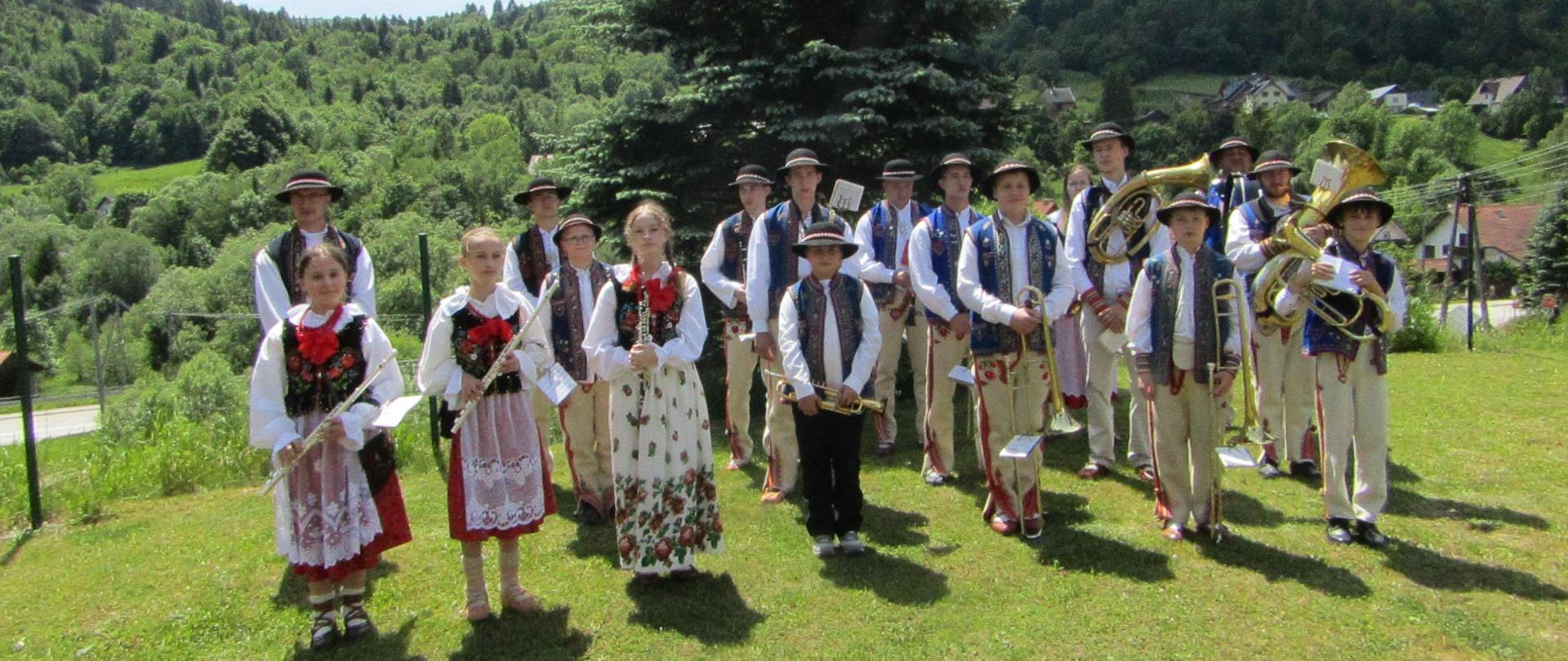 Orkiestra złożona z dzieci stoi na łące. Dzieci ubrane po góralsku w rękach trzymają instrumenty muzyczne. W tle lasy.