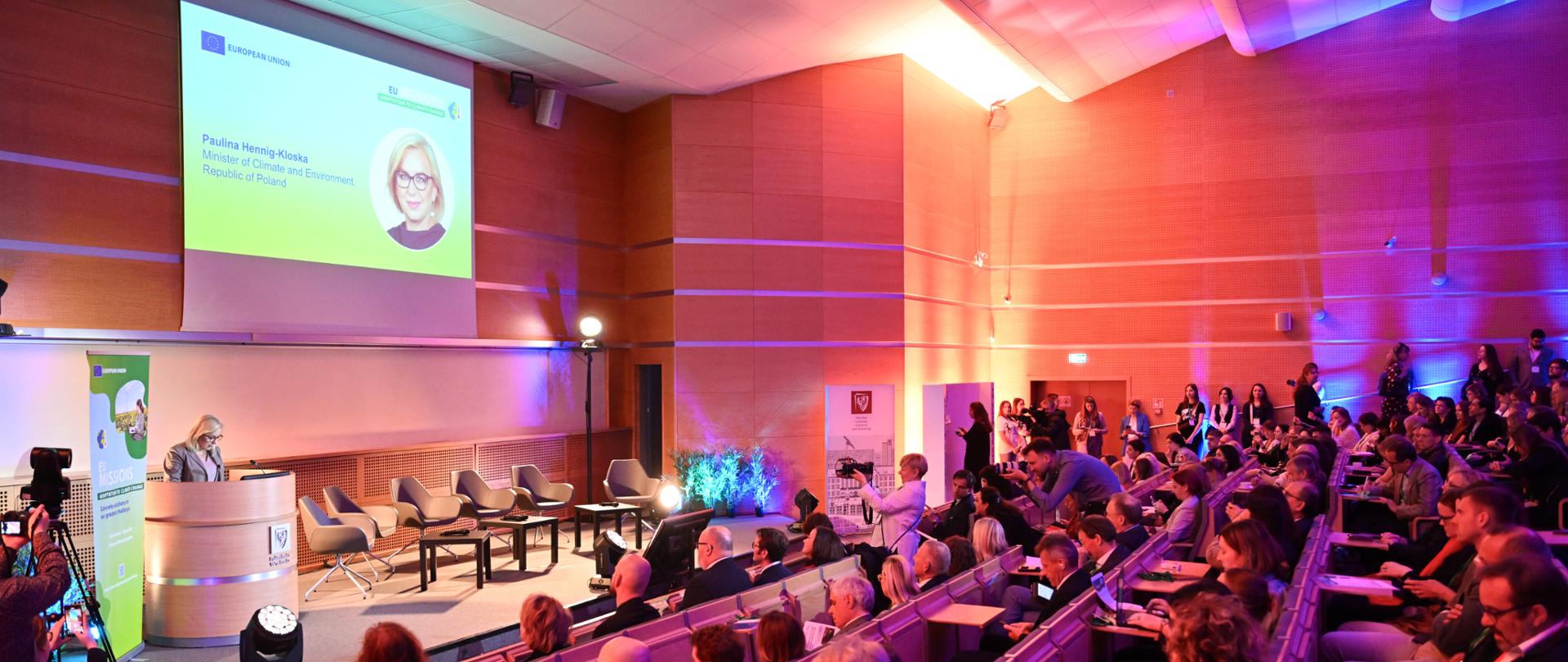  Forum Misji UE "Adaptacja do zmian klimatu" widok na salę od strony widowni i z boku scenę, na której za mównicą przemawia Paulina Hennig-Kloska