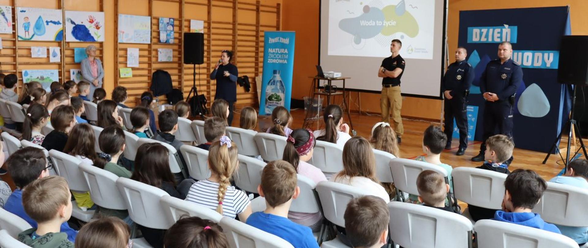 Zdjęcie przedstawia policjantów i strażaka podczas warsztatów interaktywnych dla dzieci i pracowników szkół z okazji Światowego Dnia Wody. Poruszono tematykę związaną z akcją "STOP POŻAROM TRAW" oraz bezpieczeństwa na wodzie. Policjanci i strażak w umundurowaniu stoją na tle ściany i ekranu multimedialnego, na którym wyświetlana jest prezentacja. Dzieci siedzą na krzesełkach, zwrócone w stronę osób prowadzących spotkanie.