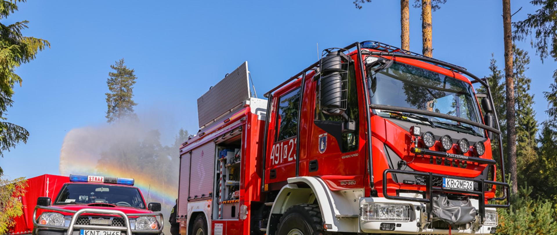 na zdjęciu są dwa czerwone samochody strażackie na polanie leśnej. w tle majaczą świerki, a za samochodami utworzyła się tęcze na tle lasu. zdjęcie zrobione zostało podczas pięknej słonecznej pogody