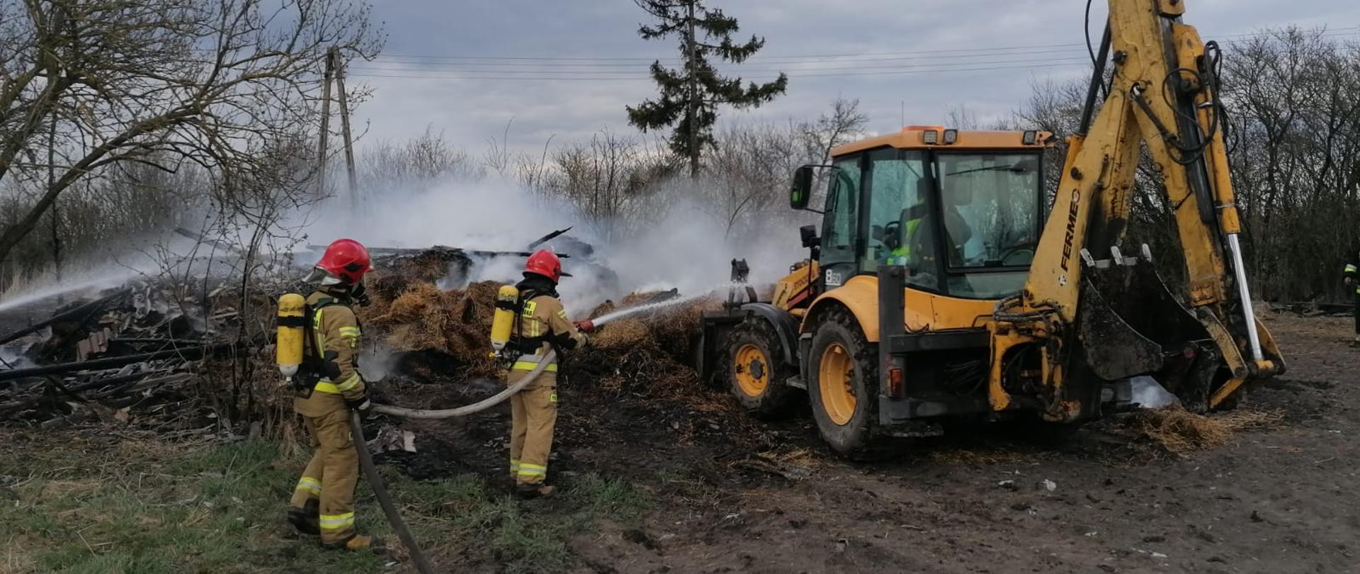 Zdjęcie przedstawia dwóch strażaków w ubraniach specjalnych z założonymi na plecach aparatami ochrony dróg oddechowych. Strażacy za pomocą linii gaśniczej prowadzą działania zmierzające do ugaszenia tlącej się słomy. Po prawej stronie widnieje koparko-ładowarka, która pomaga przerzucać zgliszcza pożarowe. 