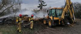 Zdjęcie przedstawia dwóch strażaków w ubraniach specjalnych z założonymi na plecach aparatami ochrony dróg oddechowych. Strażacy za pomocą linii gaśniczej prowadzą działania zmierzające do ugaszenia tlącej się słomy. Po prawej stronie widnieje koparko-ładowarka, która pomaga przerzucać zgliszcza pożarowe. 