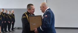 Komendant wojewódzki Policji w Gdańsku wręcza wyróżnienie strażakowi Państwowej Straży Pożarnej obok leżą wyróżnienia niedaleko stoją strażacy w umundurowaniu wyjściowym ze sznurem.