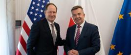 Minister Wieczorek stoi obok mężczyzny w garniturze, za nimi pod białą ścianą flagi Polski, UE i USA.