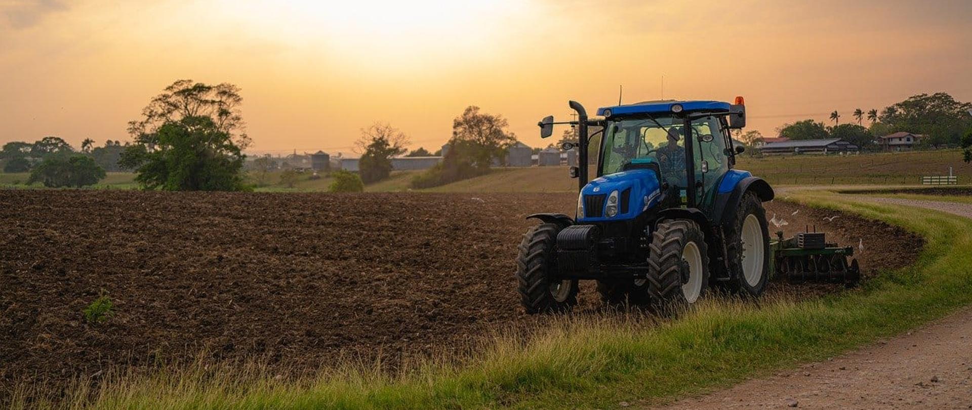Zdjęcie przedstawia nowoczesny ciągnik rolniczy marki New Holland podczas pracy na świeżo zaoranym polu. Operator wykonuje zabieg uprawowy – prawdopodobnie bronowanie lub kultywację. Ujęcie wykonane o zachodzie słońca oddaje klimat pracy rolnika na wsi. Tego typu sprzęt jest powszechnie wykorzystywany w praktycznej nauce zawodu technik rolnik lub mechanik-operator pojazdów i maszyn rolniczych.