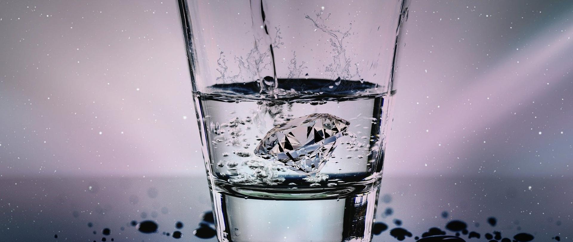 Na zdjęciu znajduje się szklanka z wodą, do której do środka wrzucony został diament. Do szklanki z góry dolewana jest woda. Wokół szklanki na szklanym blacie są krople rozlanej wody.