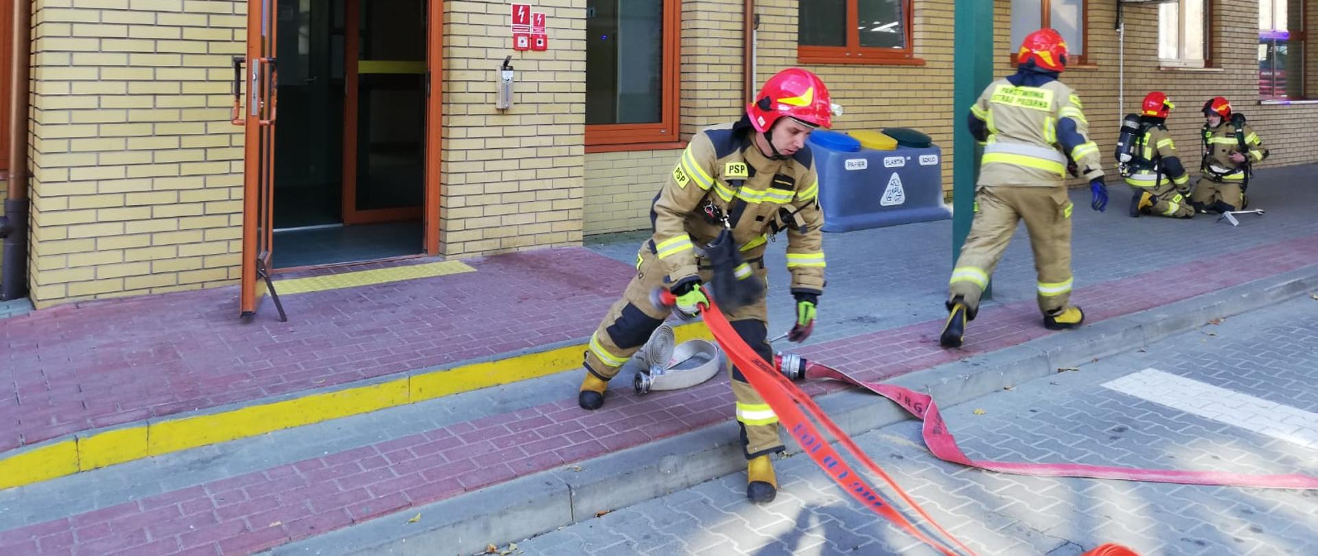 Na zdjęciu widać strażaków w ubraniach specjalnych którzy rozwijają węże pożarnicze oraz ubierają się w sprzęt ochrony dróg oddechowych przy budynku placówki straży granicznej. 