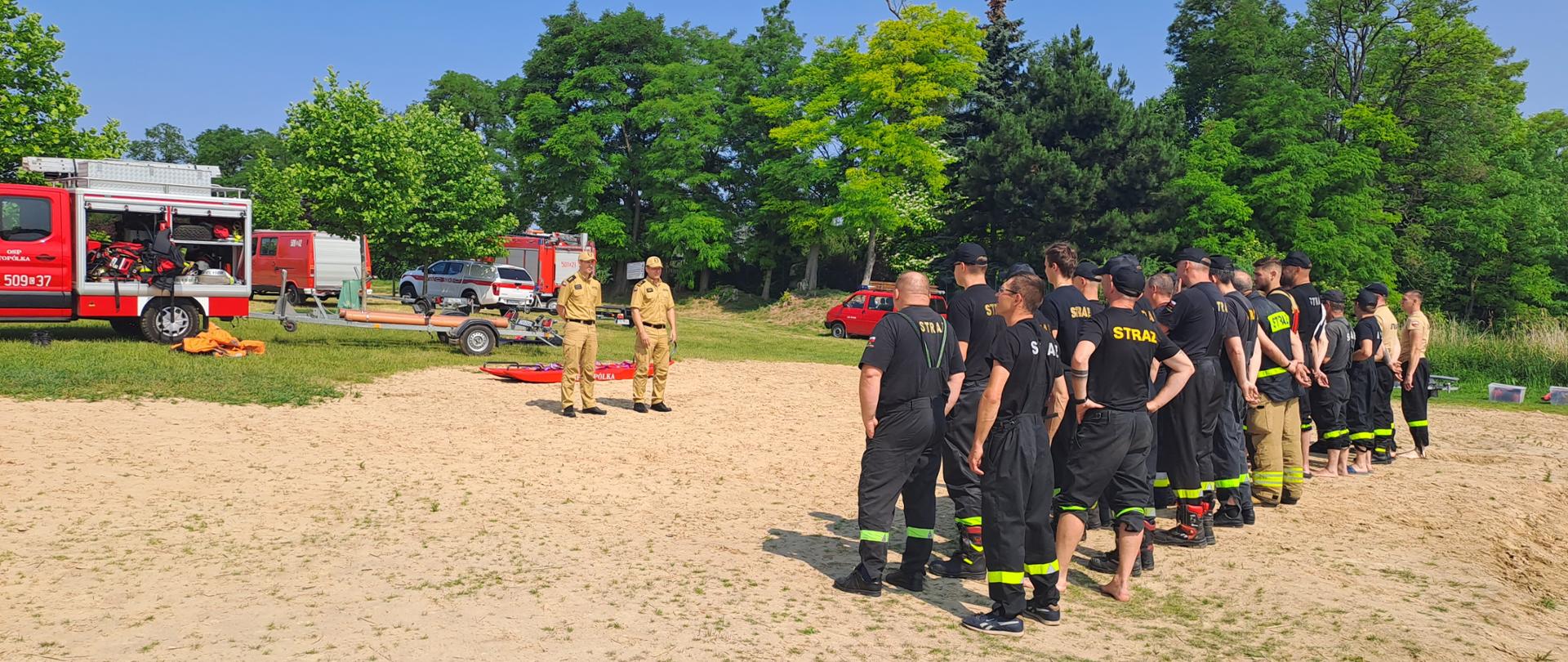 Zdjęcie przedstawia plażę, przy jeziorze głuszyńskim, na którym odbywa się odprawa strażaków PSP i OSP przed rozpoczęciem ćwiczeń. W tle samochody pożarnicze.