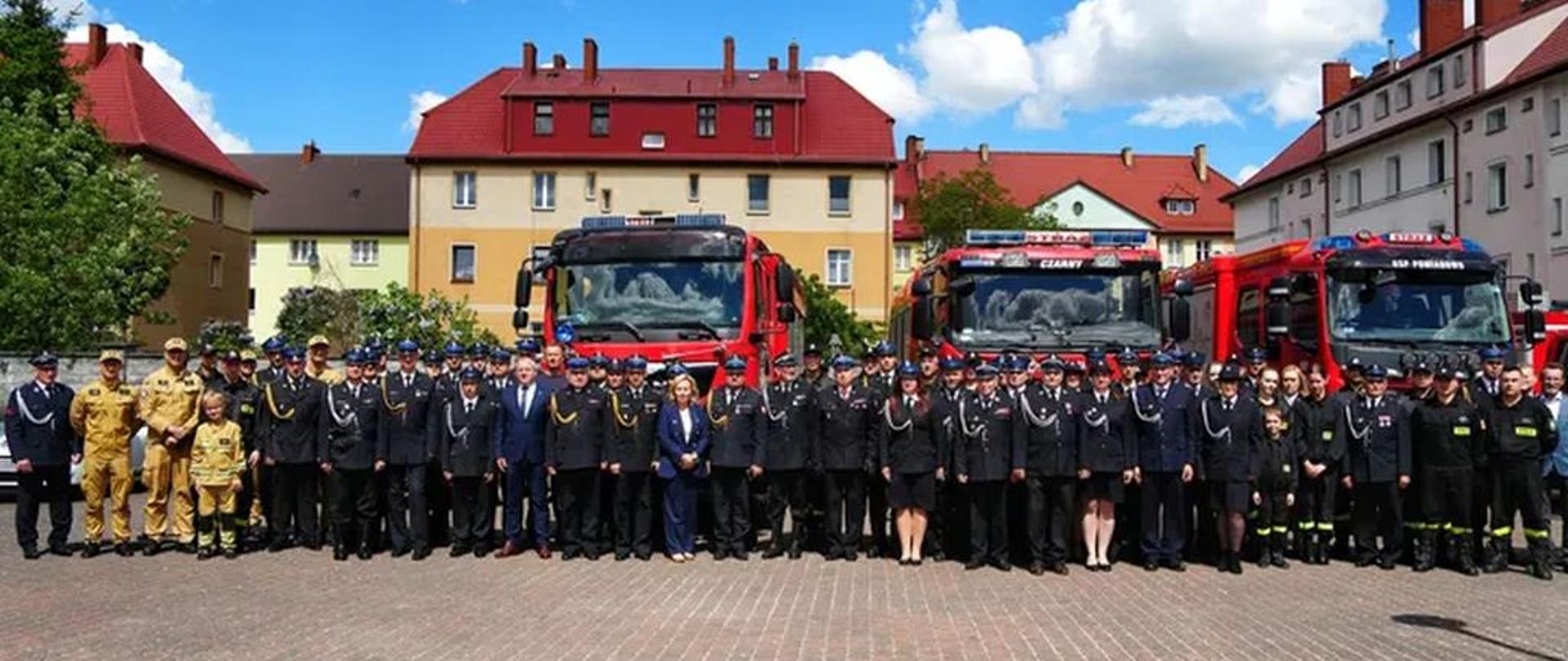 Grupowe zdjęcie uczestników uroczystości na wybrukowanym placu miejskim, wykonane w słoneczny dzień pod błękitnym niebem z białymi chmurami. W tle stoją zaparkowane trzy czerwone samochody pożarnicze. Środkowy pojazd ma na dachu napis „STRAŻ”, a na pojeździe po prawej stronie widnieje napis „OSP POMIANOWO”. Przed samochodami, w jednym długim rzędzie, stoi kilkadziesiąt osób. Większość stanowią strażacy w ciemnogranatowych mundurach wyjściowych z czapkami. Po lewej stronie rzędu stoi kilku strażaków w piaskowych mundurach bojowych, a także dwójka dzieci. W centrum oraz po prawej stronie rzędu stoją cywile – w tym kobieta w niebieskim garniturze oraz mężczyzna w ciemnej marynarce i jasnych spodniach. W tle widoczne są wielorodzinne budynki mieszkalne o jasnych elewacjach i spadzistych, czerwonych dachach.