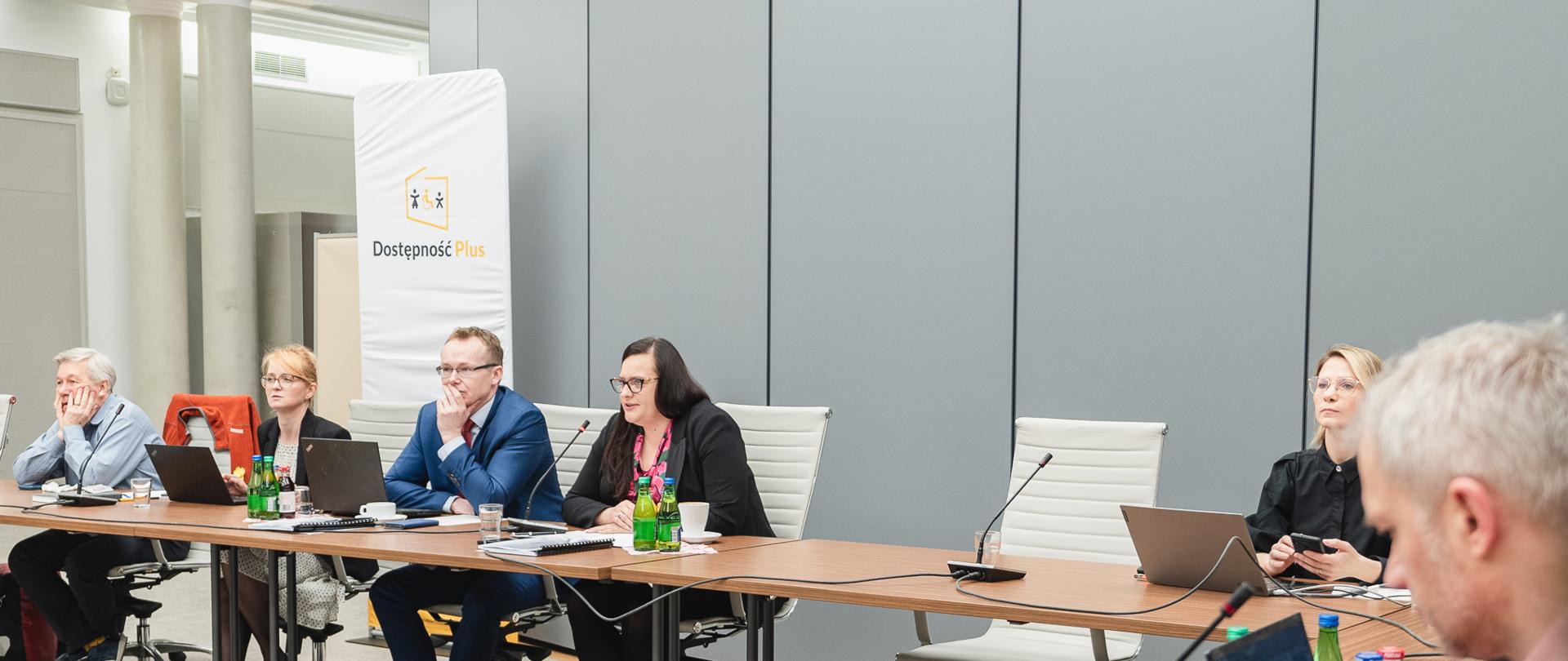 Kilkanaście osób siedzi przy dwóch prostopadle ustawionych do siebie stołach. Przy pierwszym stole w środku siedzi wiceminister funduszy i polityki regionalnej Małgorzata Jarosińska-Jedynak.