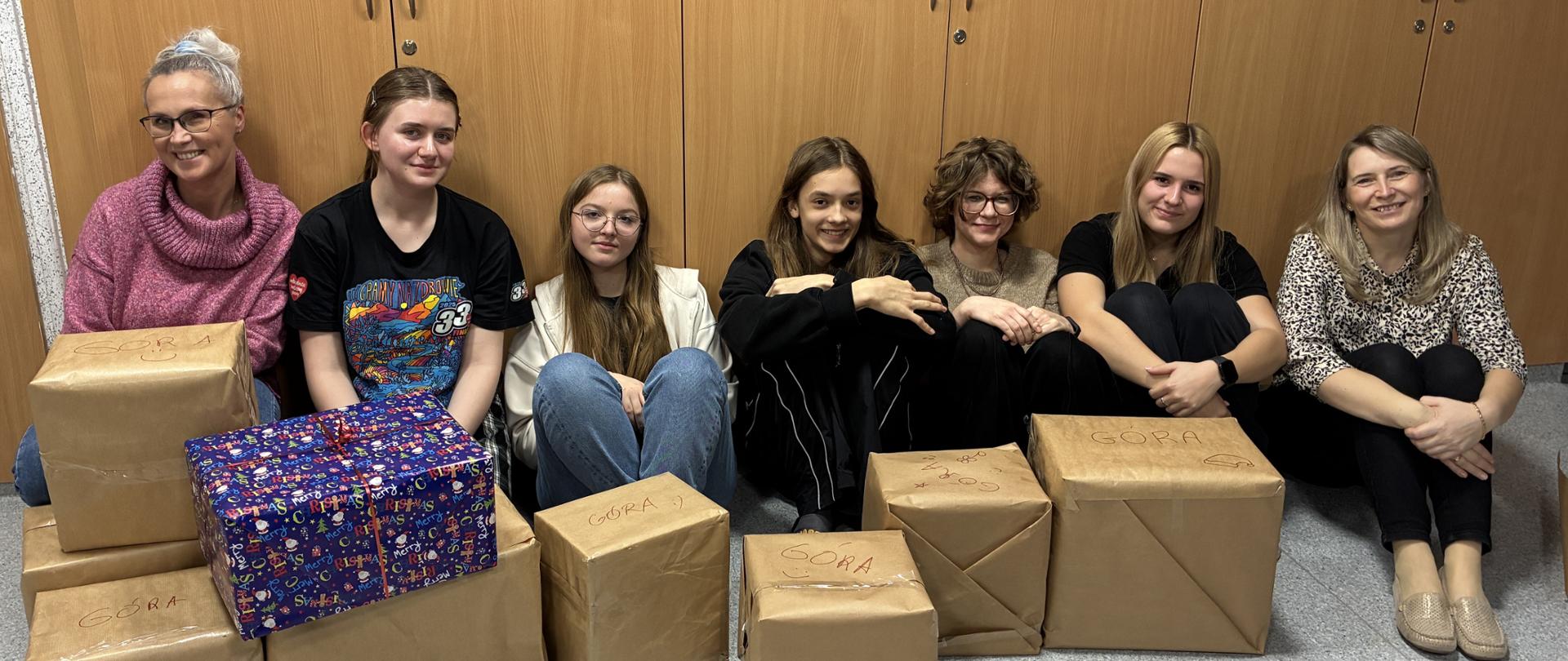 Na tle drewnianych szaf siedzi pięcioro młodych ludzi, a po bokach dwie kobiety. Przed nimi są zapakowane paczki, wszyscy uśmiechają się